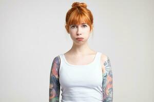 Upset young pretty redhead tattooed lady with natural makeup puffing out her cheeks while looking tiredly at camera, standing against white background photo