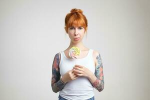 Unhappy young attractive redhead woman with tattoos holding stick candy and folding lips while looking sadly at camera, standing over white background photo