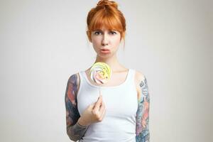 Offended young pretty redhead lady with tattoos keeping candy on stick and looking sadly at camera while standing over white background in white shirt photo