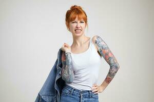 Horizontal shot of young pretty redhead lady with tattoos holding jeans coat with raised hand and frowning her face while looking at camera, isolated over white background photo