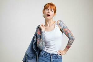 Indoor shot of attractive cocky young tattooed woman with foxy hair looking self-confidently at camera while standing against white background in casual wear photo