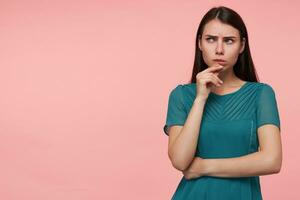 Teenage girl, serious looking woman with brunette long hair. Folding hands on a chest and touching her chin. Watching to the left at copy space over pastel pink background. Wearing emerald dress photo