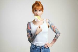 Excited young green-eyed redhead woman with tattoos keeping stick with candy in raised hand and looking surprisedly at camera, isolated over white background photo