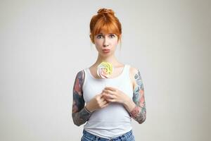 Indoor shot of young lovely redhead tattooed woman with bun hairstyle pouting her lips while looking excitedly at camera, being afraid that someone will take her candy, isolated over white background photo