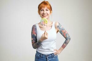Studio shot of young happy redhead tattooed lady looking cheerfully at camera with wide smile while holding candy on stick, isolated over white background photo