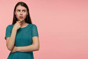 Portrait of attractive, thinking girl with long brunette hair. Folding hands on a chest and touching her chin. Watching to the right at copy space over pastel pink background. Wearing emerald dress photo