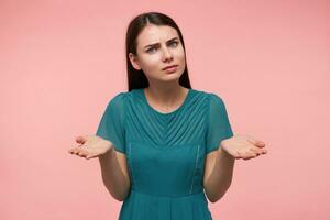 Portrait of attractive, nice looking girl with long brunette hair. Watching at the camera and holding her hands with palms up, copy space, isolated over pastel pink background. Wearing emerald dress photo