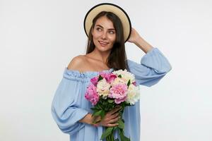 Portrait of attractive girl with long brunette hair. Wearing a hat and blue pretty dress. Holding a bouquet of flowers and touching a hat. Watching to the left at copy space over white background photo