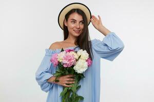 Young lady, pretty woman with long brunette hair. Wearing a hat and blue dress. Holding a bouquet of beautiful flowers and touching her hat. Watching to the right at copy space over white background photo