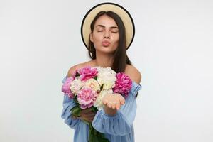 Portrait of attractive, nice looking girl with long brunette hair. Wearing a hat and blue dress. Holding a bouquet of flowers and sending a kiss. Stand isolated over white background photo