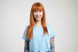 Cheerful young long haired redhead lady with tattoos showing her white perfect teeth while smiling happily at camera, isolated against white background photo
