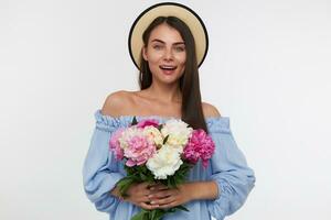 Portrait of attractive girl with big smile and long brunette hair. Wearing a hat and blue pretty dress. Holding a bouquet of beautiful flowers. Watching at the camera isolated over white background photo