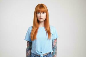 Indoor shot of bewildered young long haired redhead lady with tattoos biting her lips while looking worringly at camera, posing over white background photo