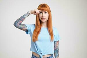 Bewildered young pretty tattooed woman with loose long hair keeping index finger on forehead and looking at it while standing over white background in blue t-shirt photo