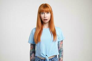 Dazed young attractive long haired redhead lady with tattoos rounding surprisedly her green eyes while looking at camera, isolated over white background photo