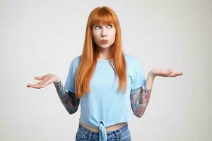 Bewildered young attractive redhead woman with tattoos pouting her lips while looking perplexedly aside and keeping palms raised, isolated over white background photo