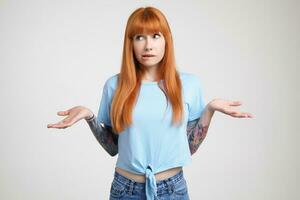Embarrassed young redhead tattooed female biting her underlip while shrugging with raised palms, dressed in casual clothes while standing over white background photo