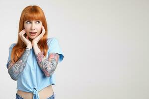 Surprised young attractive long haired redhead lady with tattoos keeping raised hands under her face while looking wonderingly aside, standing over white background photo