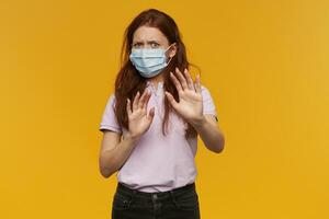 Worried scared young woman wearing medical protective mask keeps hands in front of herself and defending from threat over yellow background photo