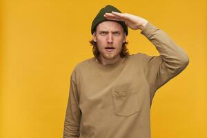 Portrait of serious male with blond hair and beard. Wearing green beanie and beige sweater. Holds palm next to forehead and gaze into distance. Watching at the camera isolated over yellow background photo