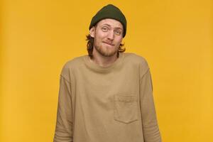 Doubtful man, unsure guy with blond hair, beard and mustache. Wearing green beanie and beige sweater. Shrugs shoulders. Don't know nothing. Watching at the camera isolated over yellow background photo