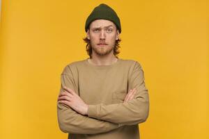 Cool looking male, bewildered bearded guy with blond hairstyle. Wearing green beanie and beige sweater. Keeps arms crossed. Watching serious at the camera isolated over yellow background photo