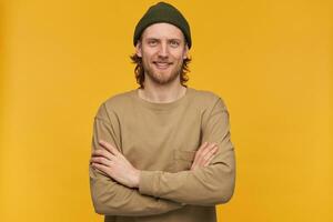 Happy man, positive guy with blond hair, beard and mustache. Wearing green beanie and beige sweater. Holds arms crossed on a chest. Watching at the camera isolated over yellow background photo