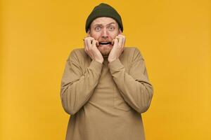 Portrait of terrified, scared male with blond hair and beard. Wearing green beanie and beige sweater. Touching his face in fear. Watching at the camera isolated over yellow background photo