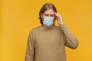 Man in pain, stressed guy with blond hair, beard. Wearing beige sweater and medical protective face mask. Touching his temple, suffer from headache. Stand isolated over yellow background photo
