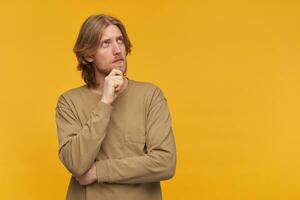 Portrait of handsome, thoughtful male with blond hairstyle and beard. Wearing beige sweater. Touching his chin. Dreamily watching to the right at copy space, isolated over yellow background photo