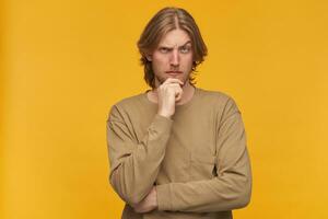 Young man, thoughtful guy with blond hair, beard and mustache. Wearing beige sweater. Touching his chin and lifts an eyebrow. Watching at the camera isolated over yellow background photo