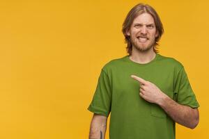 Irritated, unsure guy with blond hair, beard. Wearing green t-shirt. Has tattoo. Wry his face. Watching at the camera and pointing finger to the left at copy space, isolated over yellow background photo