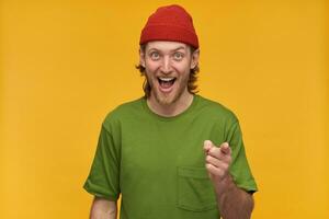 Portrait of cheerful, positive man with blond hairstyle and beard. Wearing green t-shirt and red beanie. Laughing and pointing finger at you. Watching at the camera isolated over yellow background photo
