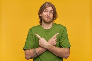 Confused, unsure bearded guy with blond hairstyle. Wearing green t-shirt. Has tattoo. Wry his face. Watching at the camera and pointing both sides at copy space, isolated over yellow background photo