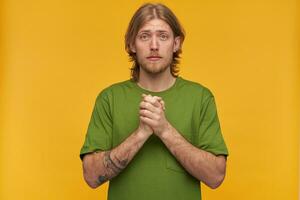 Praying male, begging bearded guy with blond hairstyle. Wearing green t-shirt. Has tattoos. Holds palms together and plead. Watching at the camera isolated over yellow background photo
