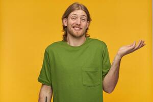 Young unsure man, cool guy with blond hair, beard and mustache. Wearing green t-shirt. Has tattoo. Shrugs and lifts his hand. So what. Watching at the camera isolated over yellow background photo