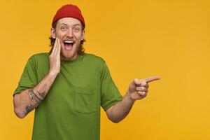 Laughing bearded guy with blond hair. Wearing green t-shirt and red beanie. Has tattoos. Watching at the camera and pointing finger to the right at copy space, isolated over yellow background photo