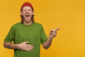 Guy with blond hair and beard. Wearing green t-shirt and red beanie. Touching belly and laughing hard with closed eyes. Pointing finger to the right at copy space, isolated over yellow background photo