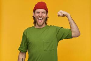 Young man, joyful guy with blond hair, beard and mustache. Wearing green t-shirt and red beanie. Has tattoo. Shows his biceps, strength. Watching at the camera isolated over yellow background photo