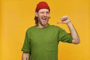 Portrait of handsome, happy male with blond hairstyle and beard. Wearing green t-shirt and red beanie. Has tattoo. Pointing thumb at himself. Winks at the camera isolated over yellow background photo