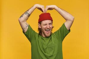 Portrait of handsome, adult male with blond hairstyle and beard. Wearing green t-shirt and red beanie. Has tattoos. Making heart sign with hands. Winks at the camera isolated over yellow background photo