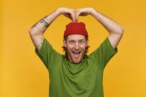 Positive, handsome bearded guy with blond hair. Wearing green t-shirt and red beanie. Has tattoos. Making heart sign with hands over his head. Watching at the camera isolated over yellow background photo