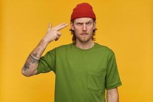 Portrait of unhappy male with blond hair and beard. Wearing green t-shirt and red beanie. Making gun gesture with fingers next to temple. Watching at the camera isolated over yellow background photo