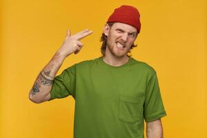 Angry bearded guy with blond hair. Wearing green t-shirt and red beanie. Has tattoos. Making gun gesture with fingers. Shoot himself in temple. Watching at the camera isolated over yellow background photo