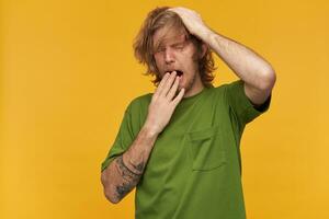 Sleepy man, tired guy with blond messy hair, beard and mustache. Wearing green t-shirt. Has tattoos. Scratch his head. Yawns covering his mouth. Stand isolated over yellow background photo