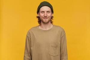 Cool looking male, handsome bearded guy with blond hair. Wearing green beanie and beige sweater. Watching at the camera with confident smile, isolated over yellow background photo