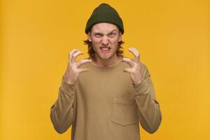 Portrait of negative, irritated male with blond hairstyle and beard. Wearing green beanie and beige sweater. Wry his face in anger. Watching at the camera isolated over yellow background photo