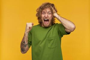 Portrait of stressed, crazy male with blond messy hairstyle and beard. Wearing green t-shirt. Has tattoos. Clench his fist. Touching head. Watching at the camera isolated over yellow background photo