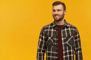 sonriente atractivo joven hombre en tartán camisa con barba en pie y mirando lejos a el lado terminado amarillo antecedentes foto
