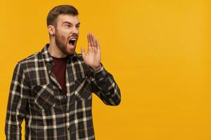 Studio shot of angry mad bearded man in plaid shirt looks aggressive looking and screaming loudly far away to the side isolated over yellow background photo